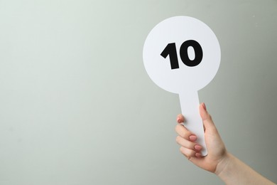 Photo of Woman holding auction paddle with number 10 on light grey background, closeup. Space for text