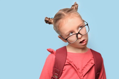 Cute little girl in glasses with backpack on light blue background. Space for text