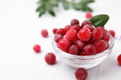 Photo of Frozen red cranberries in bowl and green leaves on white table, closeup. Space for text
