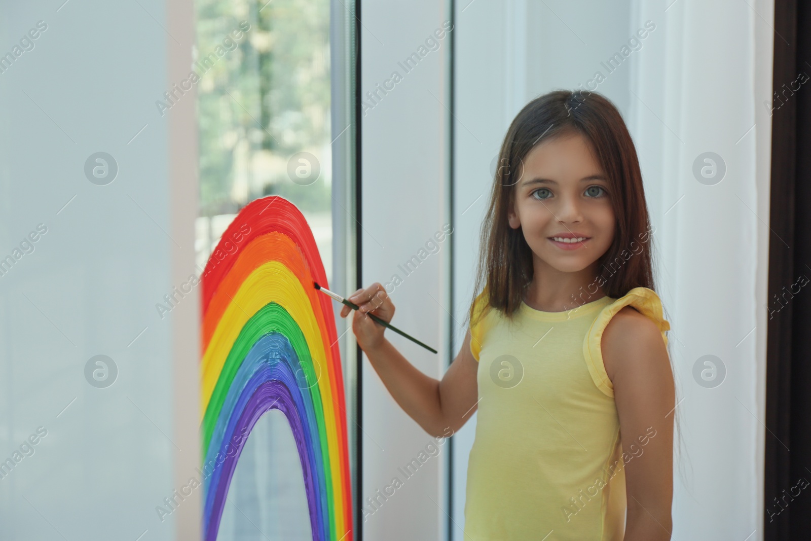 Photo of Little girl drawing rainbow on window indoors. Stay at home concept