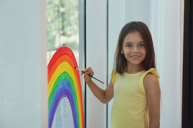 Photo of Little girl drawing rainbow on window indoors. Stay at home concept