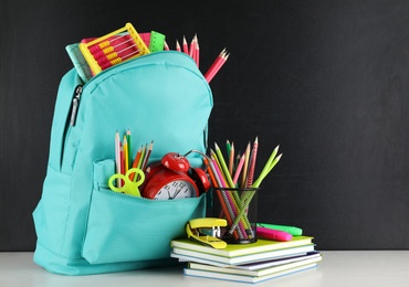 Bright backpack with school stationery on white table near black chalkboard, space for text