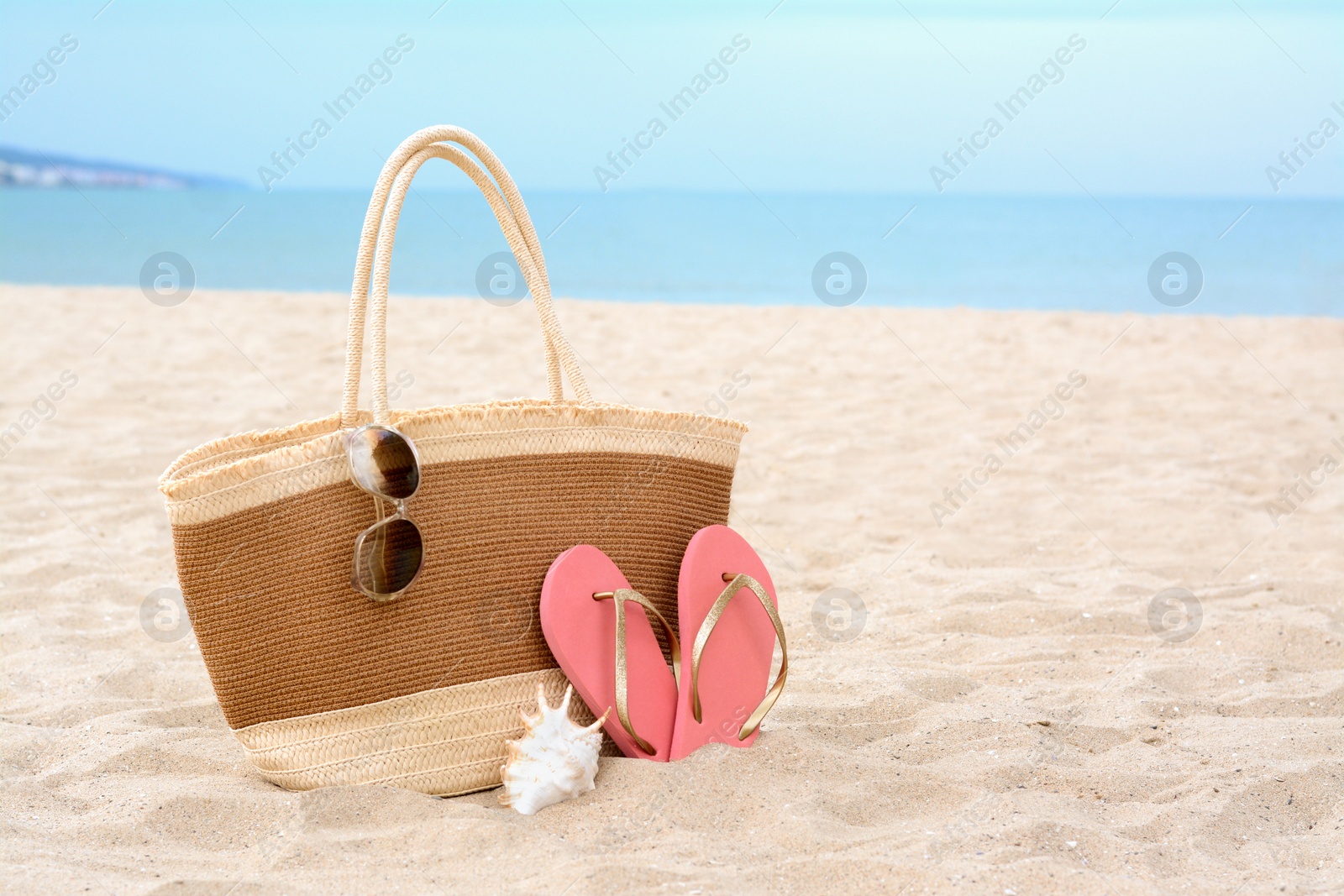 Photo of Stylish straw bag with sunglasses, flip flops and shell on sand near sea, space for text. Beach accessories