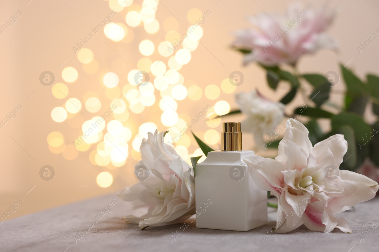 Photo of Bottle of perfume and beautiful lily flowers on table against beige background with blurred lights, closeup. Space for text