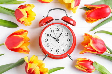 Red alarm clock, spring flowers on white wooden table, flat lay. Time change