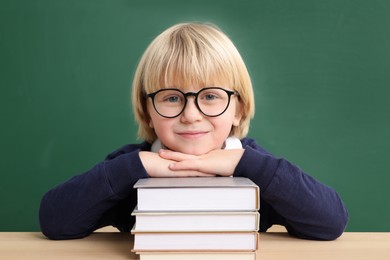 Photo of Happy little school child sitting at desk with books near chalkboard