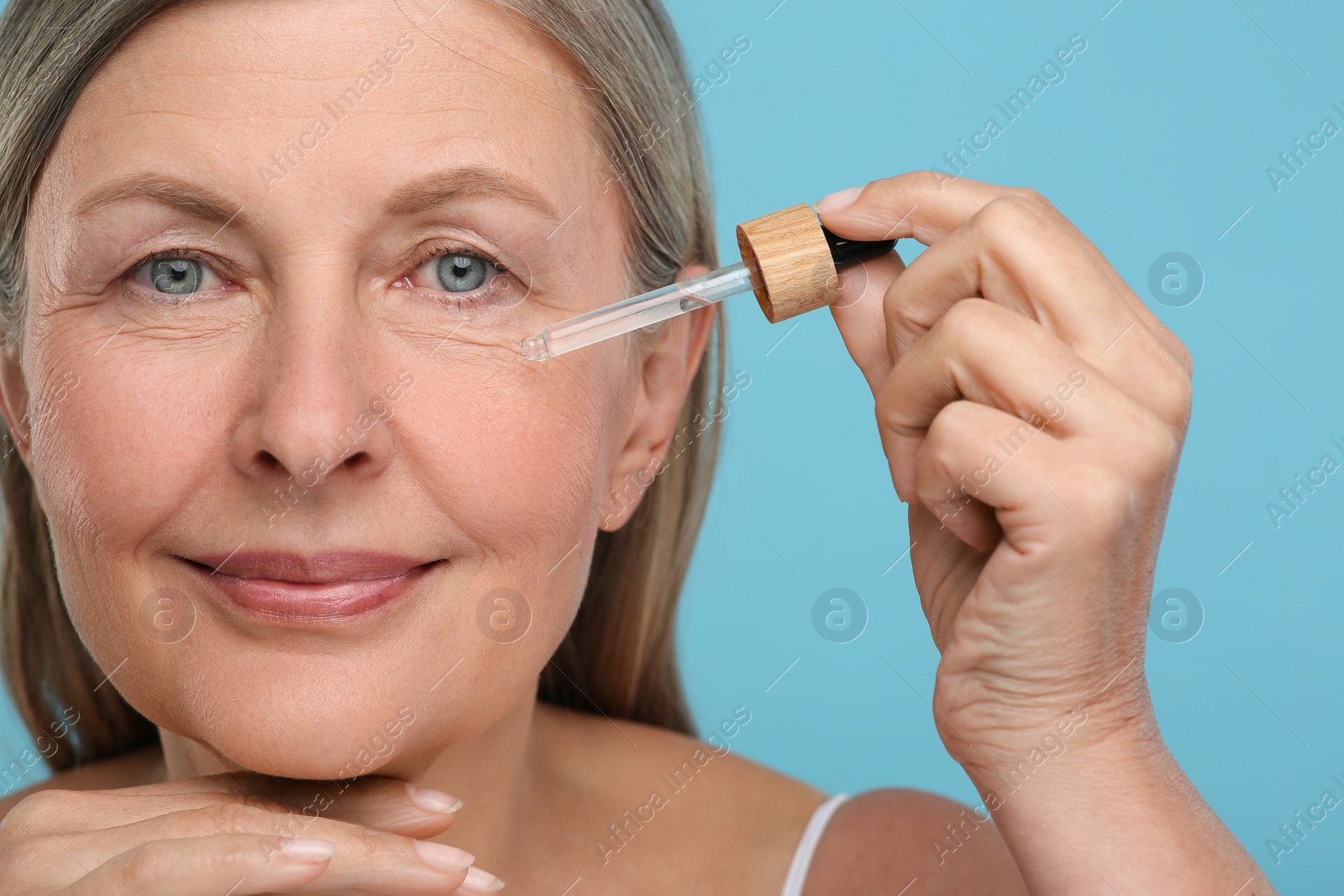 Photo of Senior woman applying cosmetic product on her aging skin against light blue background. Rejuvenation treatment