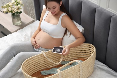 Photo of Beautiful pregnant woman with ultrasound picture of baby and basket on bed indoors