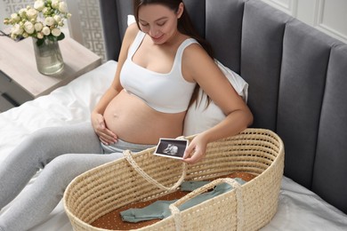 Beautiful pregnant woman with ultrasound picture of baby and basket on bed indoors