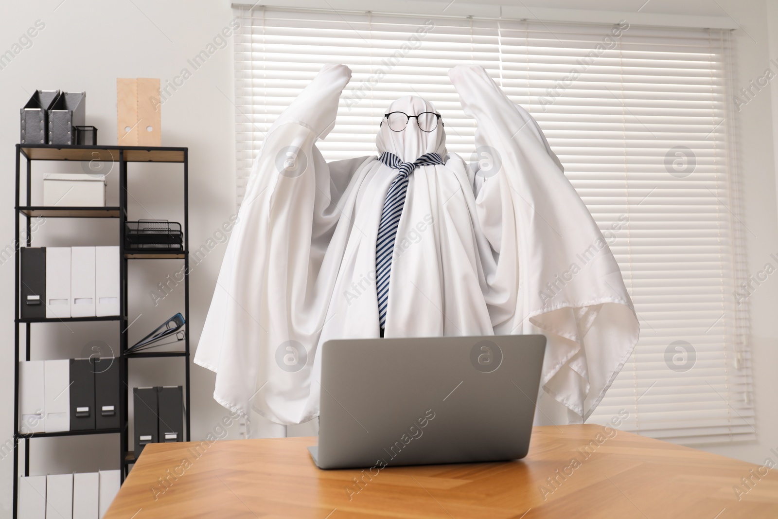 Photo of Overworked ghost. Man covered with white sheet using laptop at wooden table in office