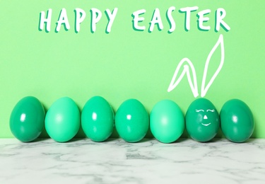 One egg with drawn face and ears as Easter bunny among others on white marble table against green background