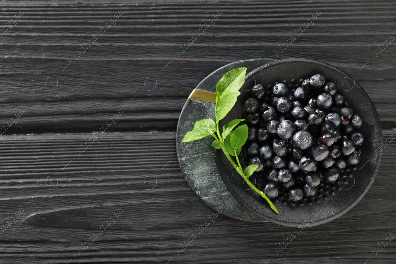 Photo of Tasty fresh bilberries with green leaves in bowl on black wooden table, top view. Space for text