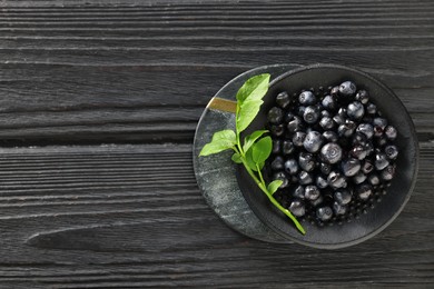 Photo of Tasty fresh bilberries with green leaves in bowl on black wooden table, top view. Space for text