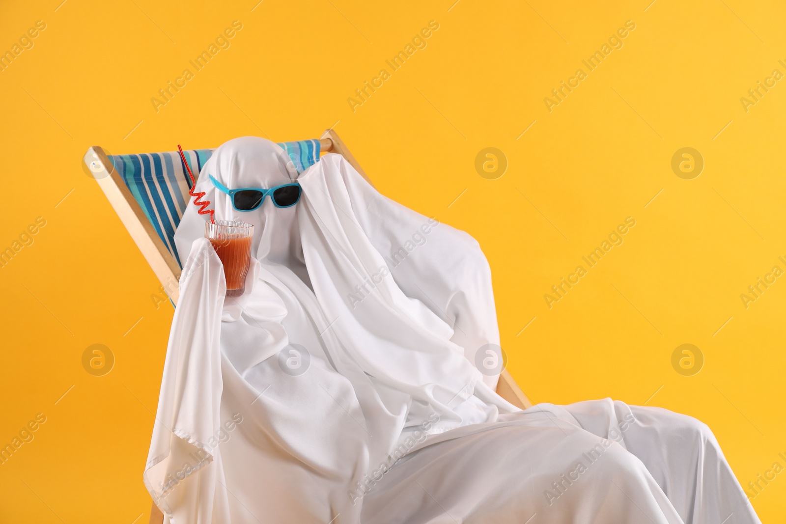 Photo of Person in ghost costume and sunglasses with glass of drink relaxing on deckchair against yellow background