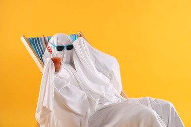 Person in ghost costume and sunglasses with glass of drink relaxing on deckchair against yellow background