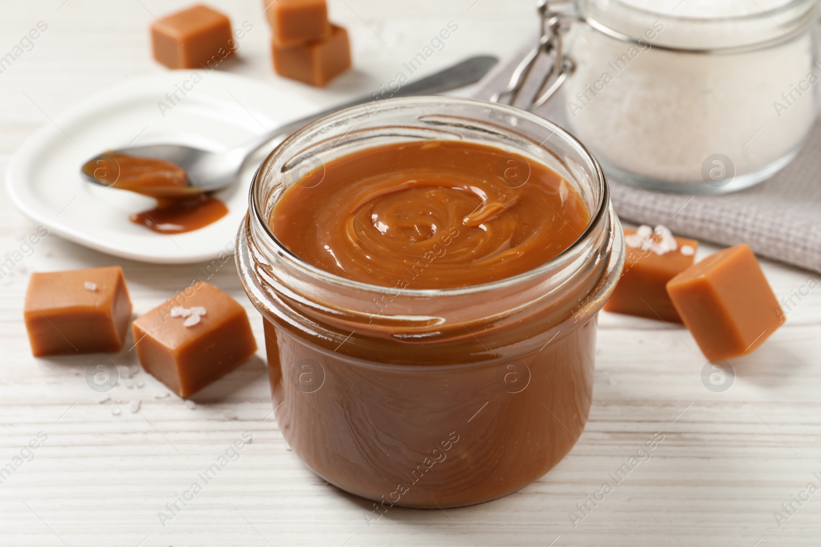 Photo of Yummy salted caramel in glass jar and candies on white wooden table, closeup