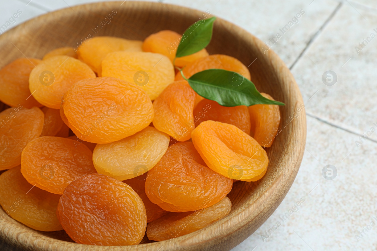 Photo of Wooden bowl of tasty apricots on white tiled table, closeup. Dried fruits