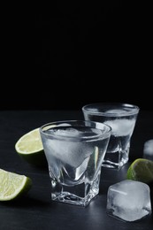 Photo of Shot glasses of vodka with lime slices and ice on black table
