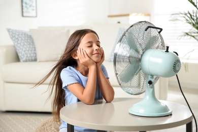 Photo of Little girl enjoying air flow from fan at home. Summer heat