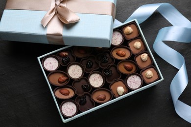 Open box of delicious chocolate candies and light blue ribbon on black table, flat lay