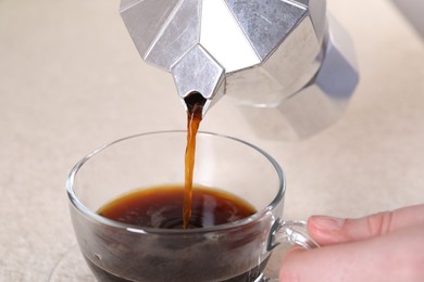 Photo of Woman pouring aromatic coffee from moka pot into glass cup at light table, closeup