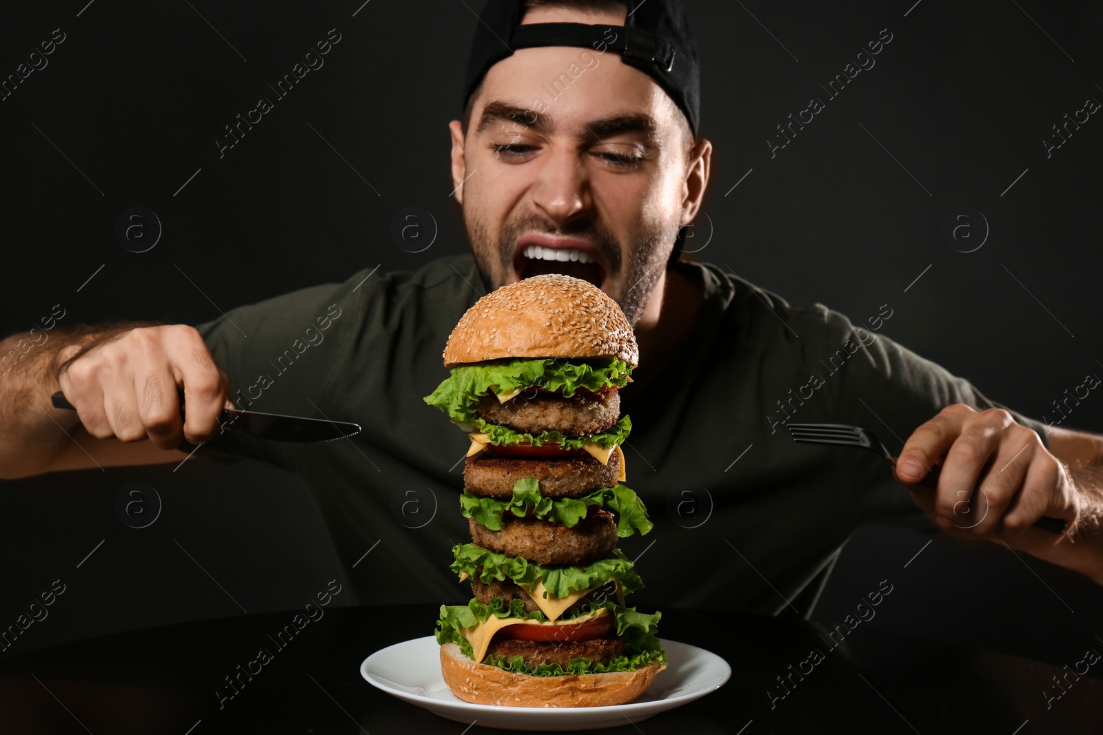 Photo of Young hungry man with cutlery eating huge burger on black background