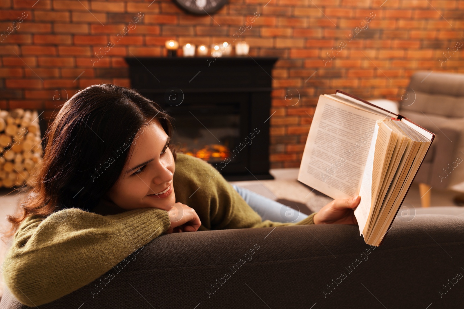 Photo of Young woman reading book on sofa near fireplace at home