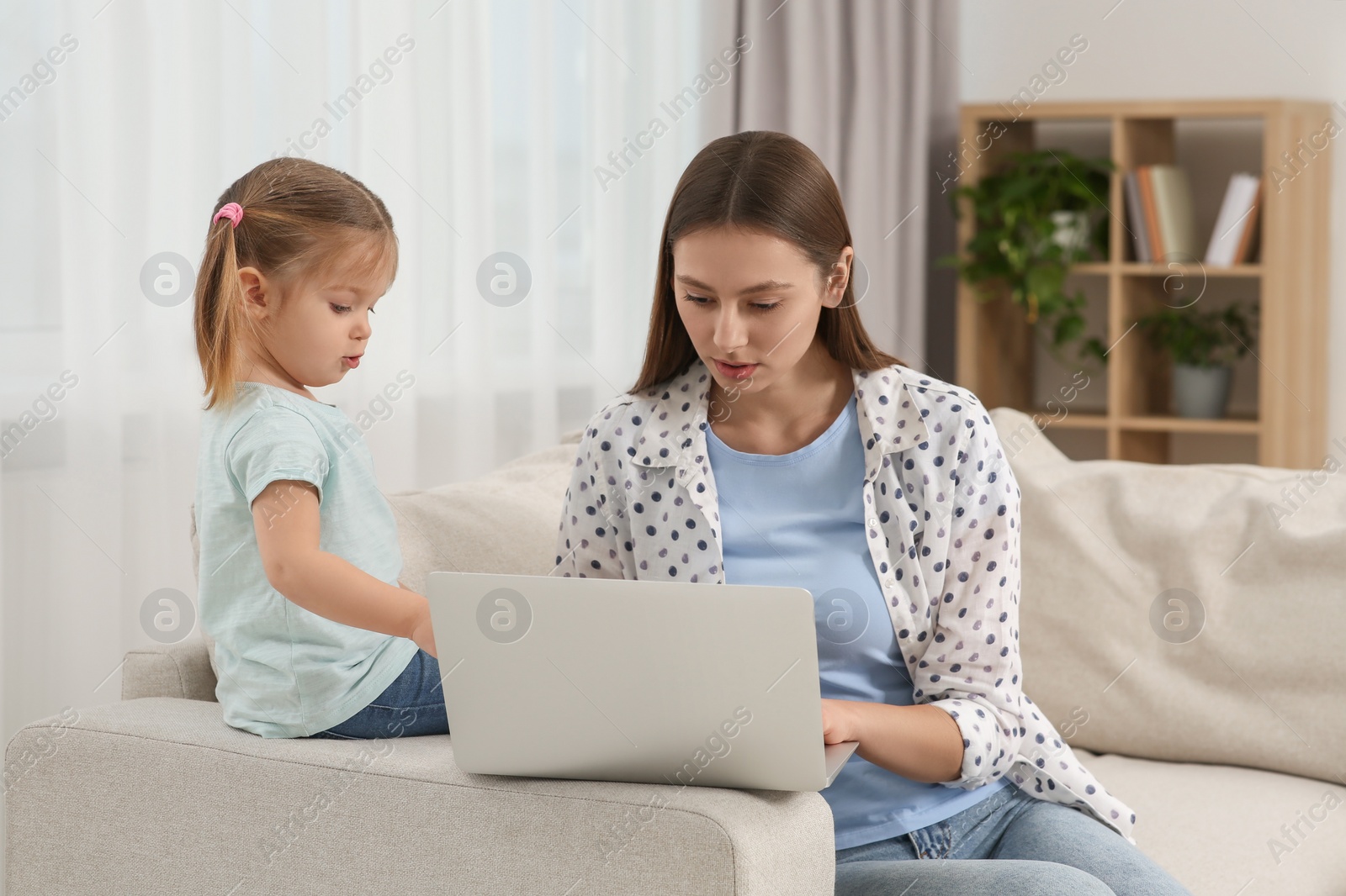 Photo of Woman working remotely at home. Little daughter bothering her mother on sofa in living room