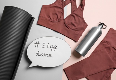 Photo of Stylish sportswear, yoga mat and speech bubble with hashtag Stay at Home on color background, flat lay. Self isolation during COVID‑19 pandemic