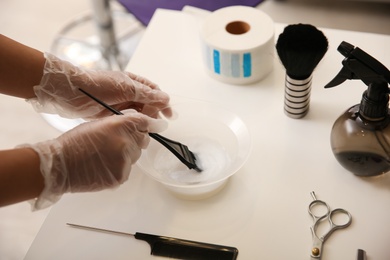 Professional hairdresser preparing dye for hair coloring at white table in beauty salon, closeup