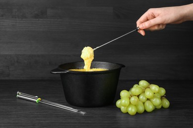 Photo of Woman dipping grape into fondue pot with melted cheese at black wooden table, closeup