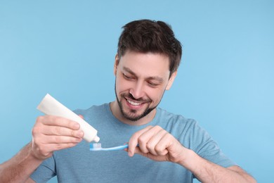 Photo of Happy man squeezing toothpaste from tube onto plastic toothbrush on light blue background