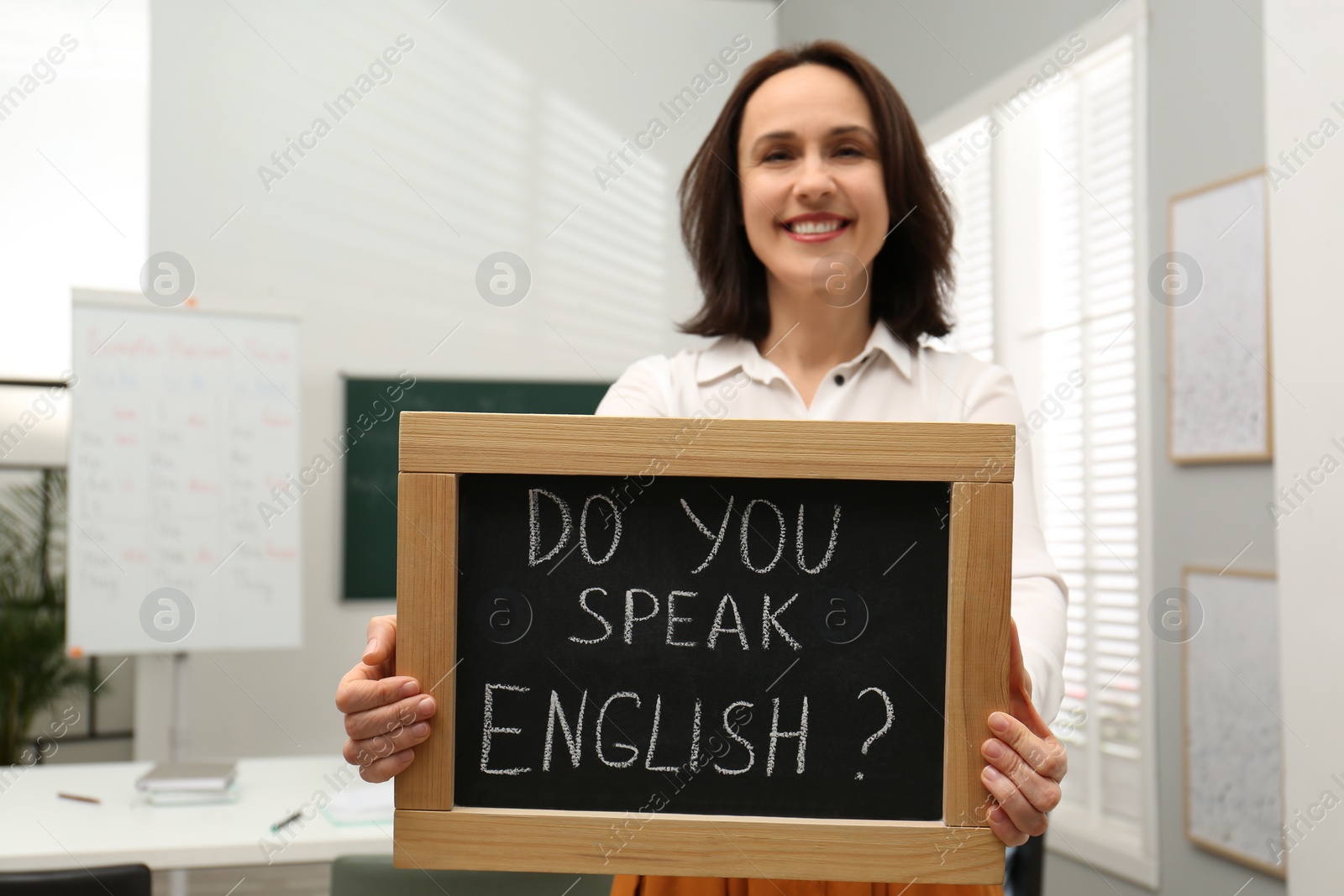 Photo of Teacher holding small chalkboard with inscription Do You Speak English? in classroom