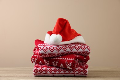 Stack of different Christmas sweaters and Santa hat on wooden table against beige background
