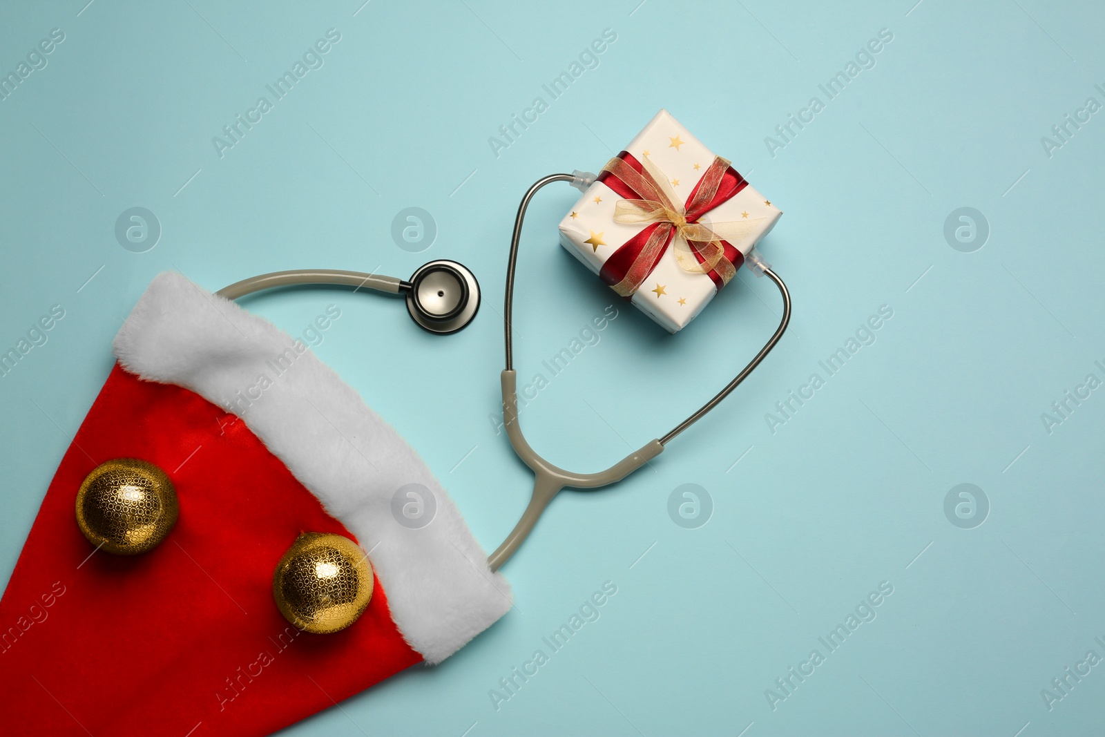 Photo of Greeting card for doctor with stethoscope, gift box, Santa hat and Christmas decor on light blue background, flat lay