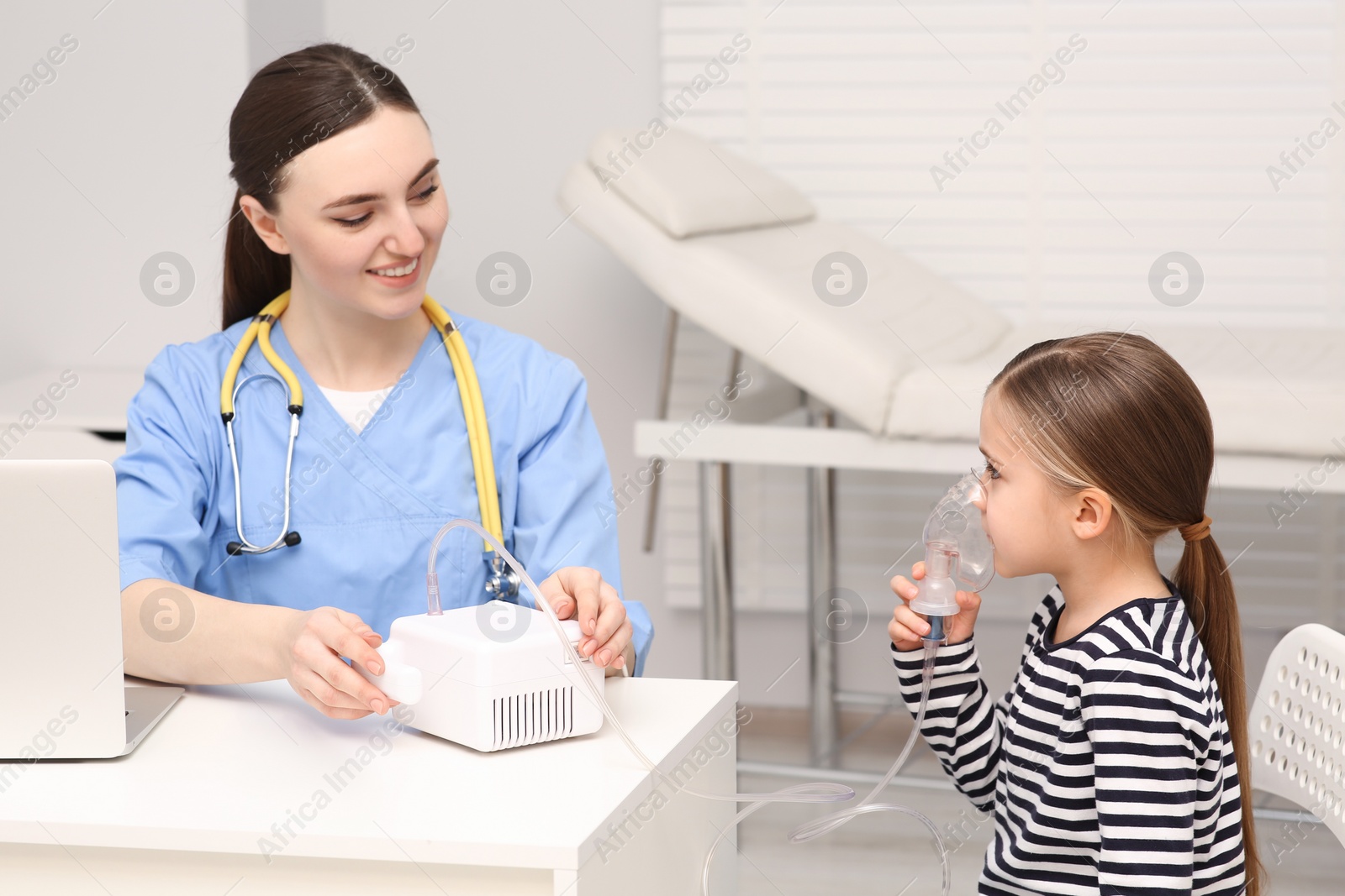 Photo of Medical assistant sitting near sick little girl while she using nebulizer for inhalation in hospital