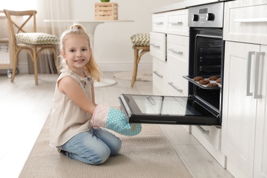 Photo of Little girl opening door of oven with cookies in kitchen