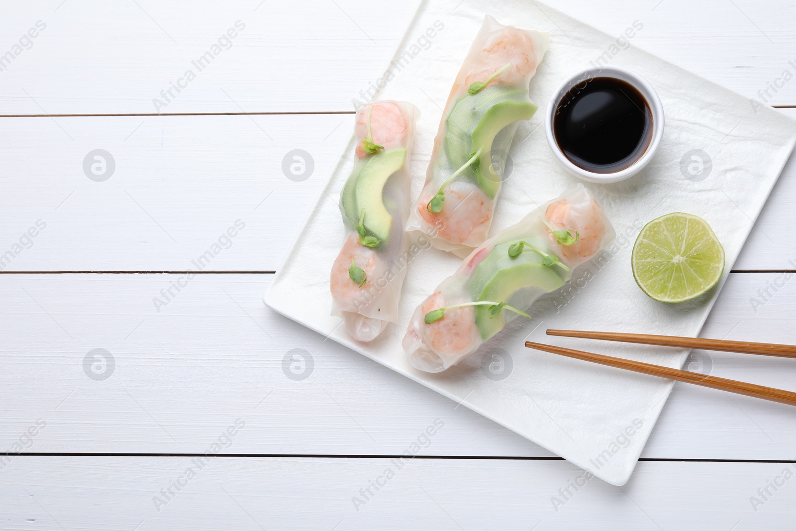 Photo of Delicious spring rolls, lime, soy sauce and chopsticks on white wooden table, top view. Space for text