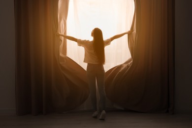 Woman opening stylish curtains at home, back view