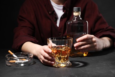 Photo of Alcohol addiction. Woman with whiskey and smoldering cigarettes at dark textured table, closeup