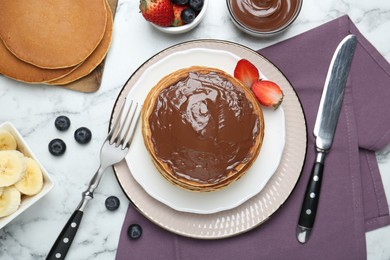 Photo of Tasty pancakes with chocolate paste, berries and cutlery on white marble table, flat lay