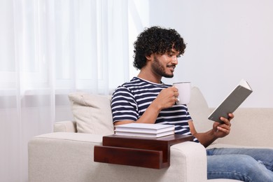 Photo of Happy man reading book and holding cup of drink on sofa with wooden armrest table at home