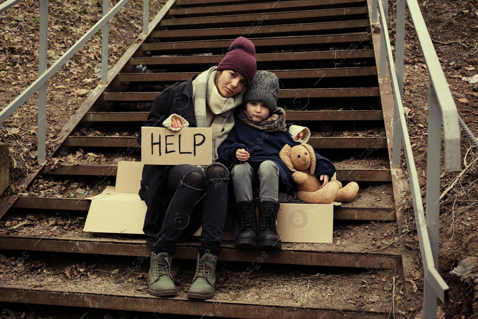 Photo of Poor mother and daughter with HELP sign sitting on stairs outdoors