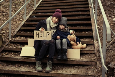 Photo of Poor mother and daughter with HELP sign sitting on stairs outdoors