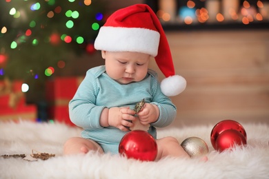 Little baby in Santa hat playing with Christmas decoration against blurred festive lights indoors