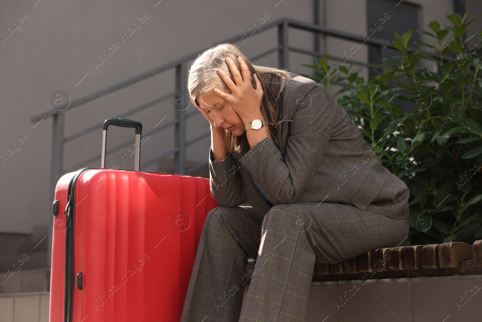 Photo of Being late. Frustrated senior businesswoman with red suitcase sitting on bench outdoors