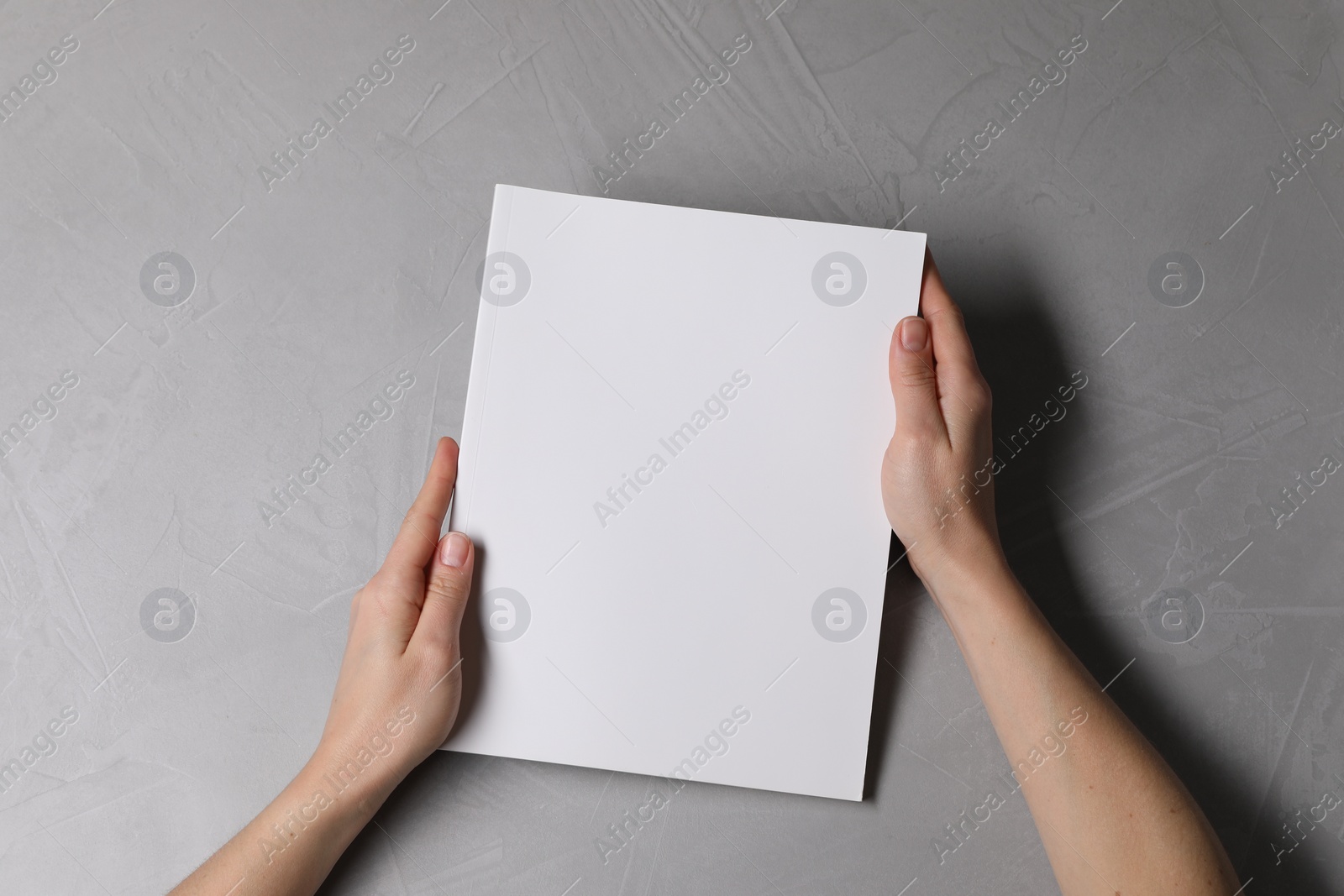 Photo of Woman holding notebook with blank cover at light grey table, top view. Mockup for design