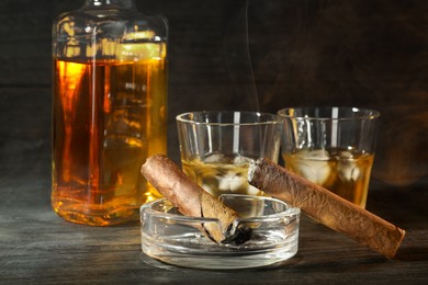 Photo of Cigars, ashtray and whiskey with ice cubes on black wooden table, closeup