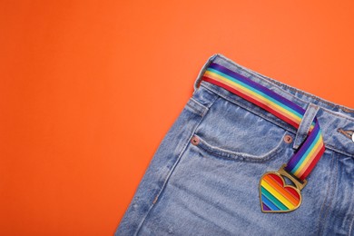 Photo of Jeans and rainbow ribbon with heart shaped pendant on orange background, top view and space for text. LGBT pride
