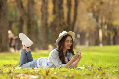 Photo of Young woman lying on green grass and using smartphone in park, space for text